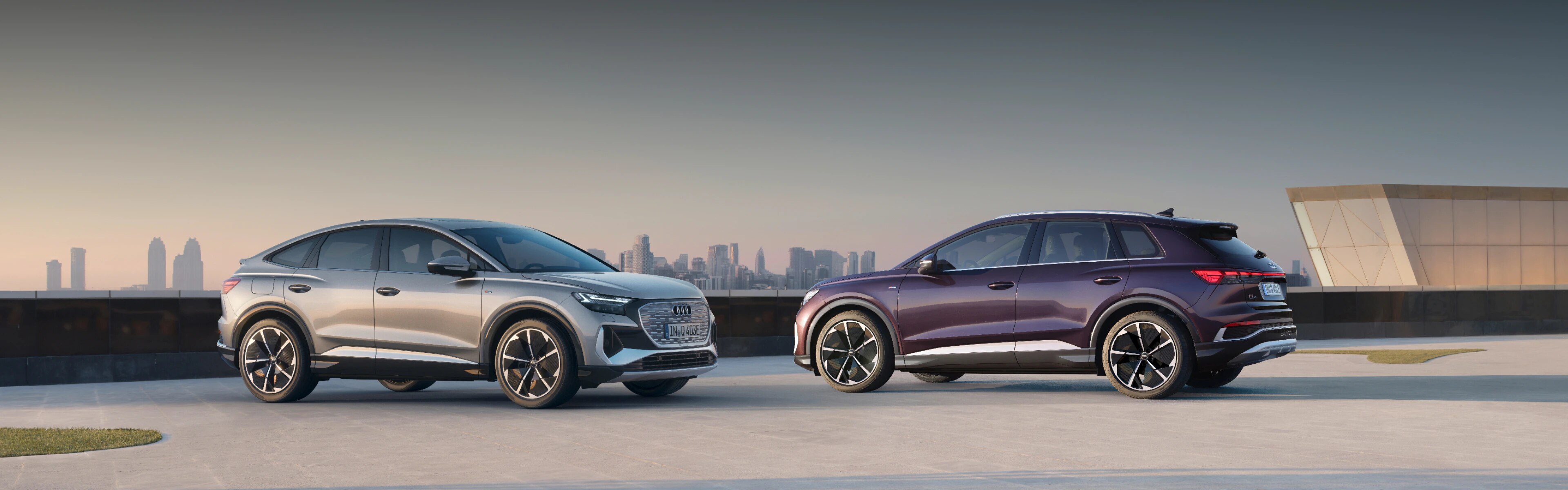 Audi Q4 e-tron Sportback and Audi Q4 e-tron parked facing each other with a city skyline in the background. 