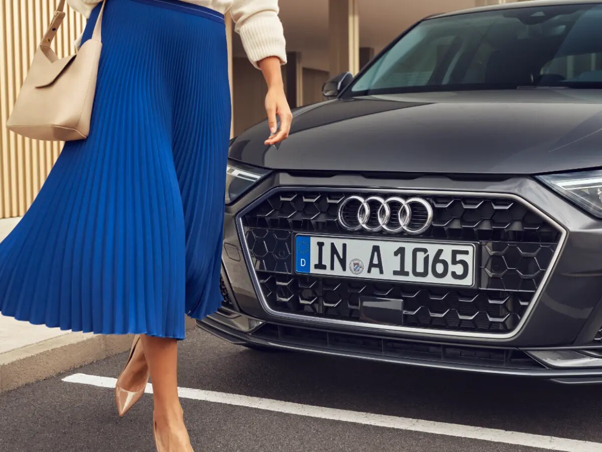 Woman from the waist below in a blue pleated skirt walking in front of an Audi. 