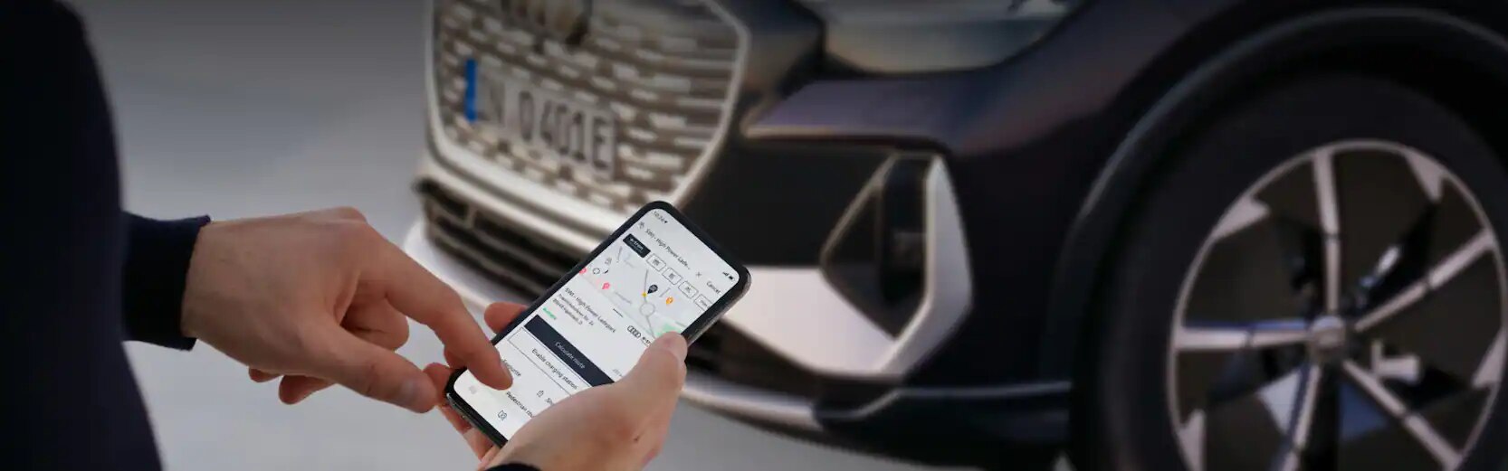 Person using the myAudi app on their smartphone to find out their route and charging stations along the way. The front of an Audi Q4 e-tron is visible in the background.