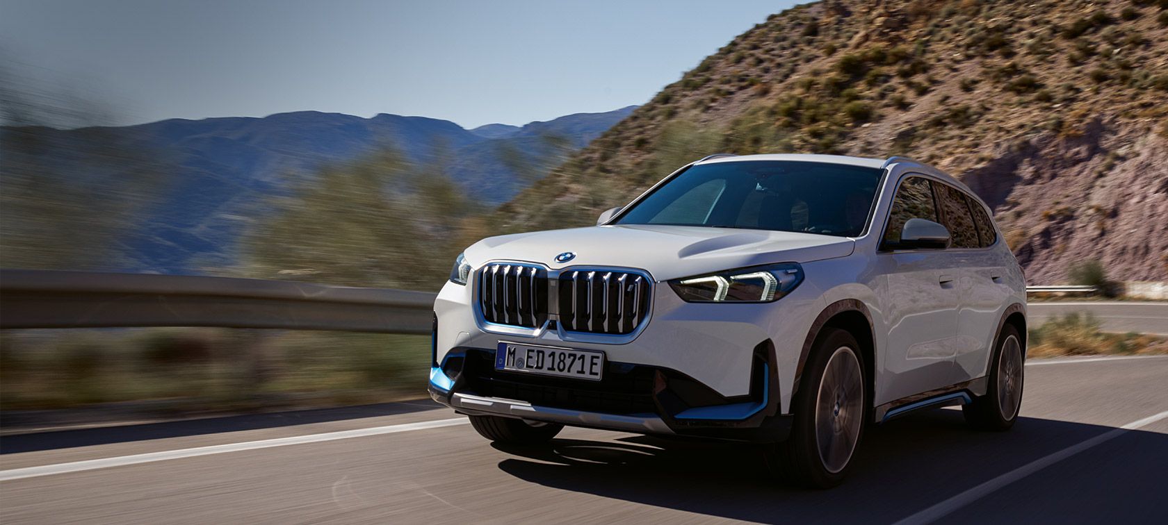 A white, all-electric BMW iX1 travelling along a cliffside road.