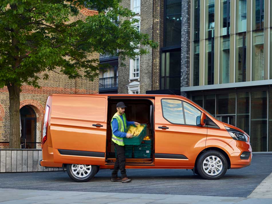 Used Ford Transit Custom Preview Image featuring a front on shot of an orange second hand Ford Transit Custom parked outside commercial offices with a delivery person removing parcels from the drivers side of the Used Ford Transit.